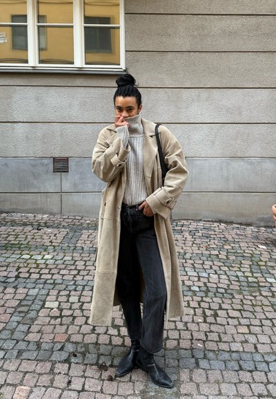 Abrigo de lana beige con mangas oversized, suéter de cuello alto gris de canalé, jeans negros y botas de tobillo, todo en un fondo de adoquines.