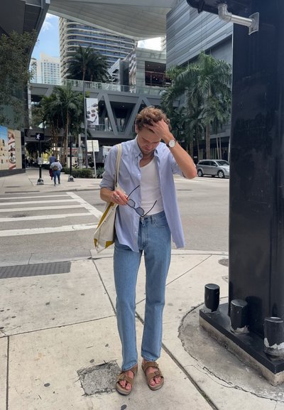 Camisa de rayas azul claro, camiseta blanca de tirantes, jeans azul claro, sandalias marrones, llevando una bolsa de tote crema y amarilla, de pie en una acera.