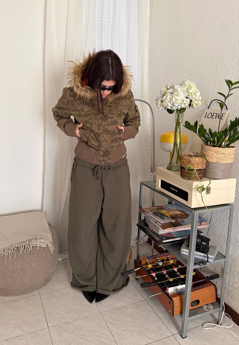 Woman wearing camouflage jacket with fur hood and wide olive pants stands indoors beside a metal shelf holding plants, magazines, and a foosball table.