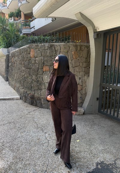 Traje de ante marrón con detalles de flecos, combinado con una blusa negra y accesorios. Fuerte contraste con el fondo de pared de piedra clara.