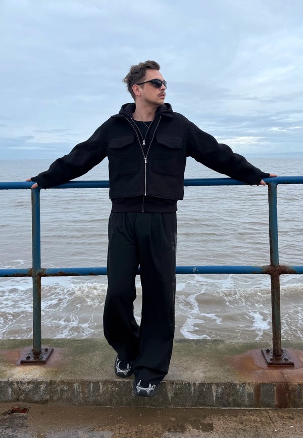 Άνδρας με μαύρο μπουφάν και γυαλιά ηλίου ακουμπάει στην μπλε μεταλλική κουπαστή δίπλα στη θάλασσα κάτω από συννεφιασμένο ουρανό.