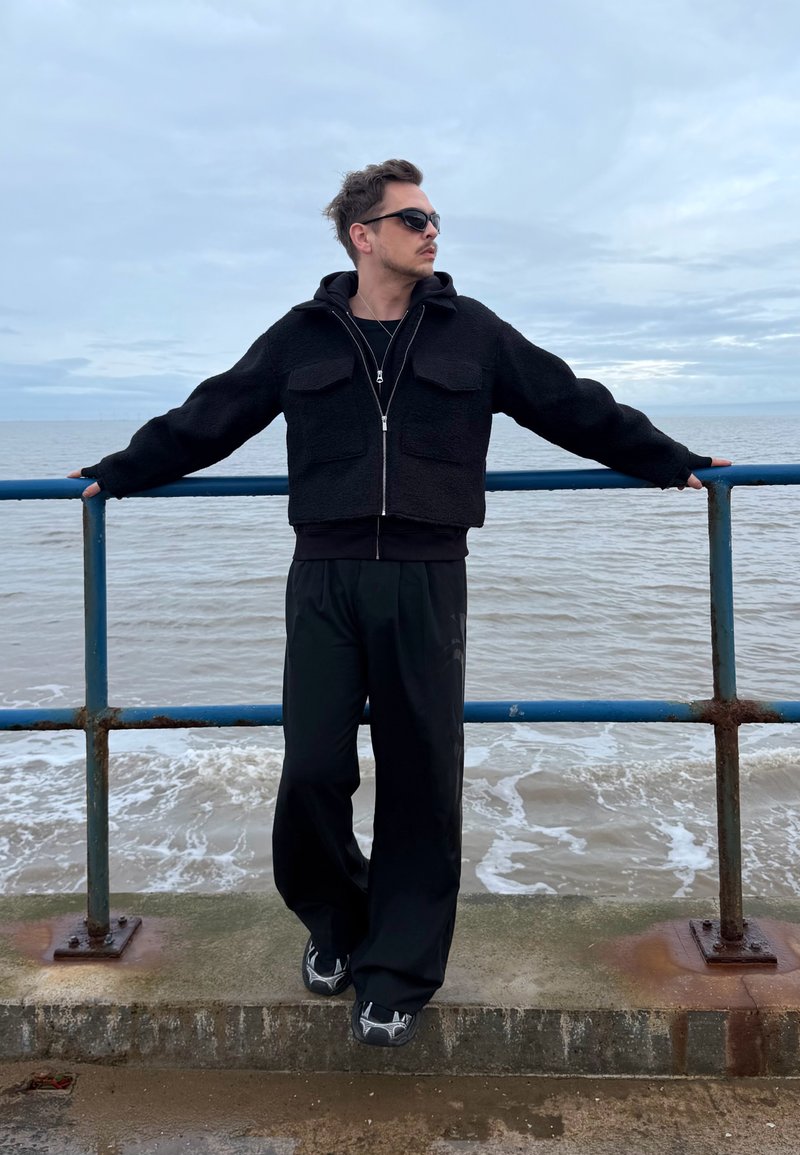 Mann iført svart jakke og solbriller lener seg mot en blå metallrekkverk ved havet under en skyet himmel.