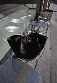 Stor mørkebrun ruskinds tote bag med en struktureret form, der har en sølvfarvet lås og to lange håndtag, placeret på en metalstol.