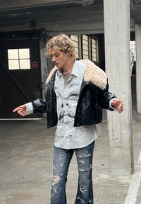 Czarna skórzana kurtka o krótkim fasonie z kołnierzem z futerka, koszula w paski z nadrukiem graficznym, podniszczone niebieskie dżinsy, stojąca w betonowej przestrzeni.