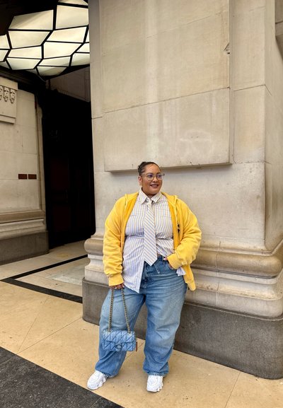 Sudadera amarilla, camisa a rayas y corbata, combinados con unos vaqueros holgados. Zapatillas blancas y un bolso estampado completan el atuendo.