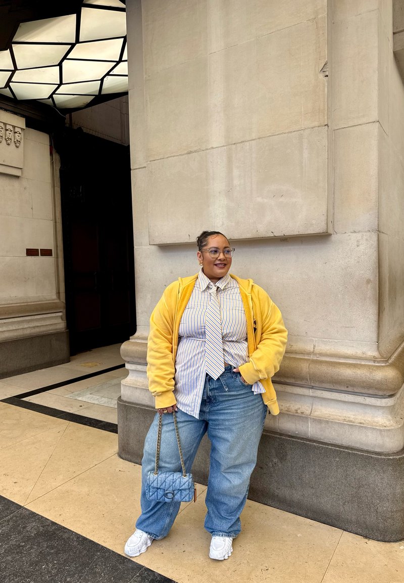 Yellow hoodie, striped shirt, and tie, paired with loose denim jeans. White sneakers and a patterned handbag complete the outfit.
