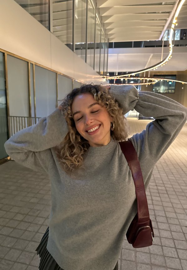 Mujer con cabello rizado y pendientes de aro sonríe con los ojos cerrados, manos detrás de la cabeza, vestida con suéter gris y bolso rojo al hombro en un espacio al aire libre iluminado.