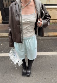 Braune Lederjacke über einem grauen Tanktop, kombiniert mit hellblauen Shorts, schwarzen knielangen Stiefeln und einer kleinen weißen Tasche.