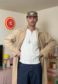 Bēšs trančs ar platajām atlokām, balta Henlija krekls un zilas džinsa bikses. Brūna raibi svītraina cepure ar baltu izšūtu burtu "C".