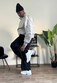 Silver bomber jacket, black wide-leg pants, white chunky sneakers, layered rings, black beanie, standing pose with one leg raised. Green plant in background.