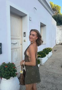 Joven mujer en un vestido oscuro sin mangas sosteniendo un bolso de flecos marrón sonríe junto a una puerta blanca con flores enmacetadas cerca.