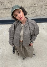 Grey bomber jacket with a textured finish, cropped beige top, and a pleated khaki skirt. Green cap with faux fur trim and a patterned bag.