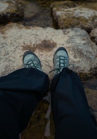 Osoba v turistických topánkach a tmavých nohaviciach stojaca na skalnatom teréne pri plytkej vode.