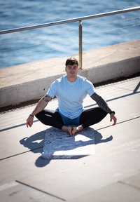 Osoba w jasnoniebieskiej koszulce z krótkim rękawem i czarnych legginsach siedzi po turecku na szarej, marmurkowej macie do jogi nad brzegiem wody, z wyrazem spokojnej twarzy.