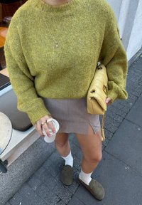 Osoba koja nosi zeleni pleteni pulover, bež suknju s prorezom, bijele čarape, cipele bez vezica, drži žutu jastučić torbicu i šalicu kave.