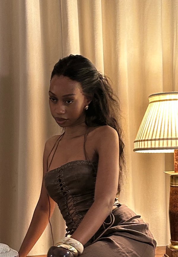 Jeune femme aux longs cheveux bruns portant un haut style corset marron et des bracelets, assise près d'une lampe allumée et de rideaux beiges à l'intérieur.