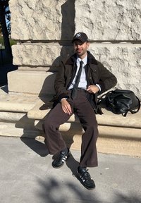 Dark green jacket, white striped shirt, black tie, brown trousers, black trainers with white stripes, sitting on a stone surface with a black bag.