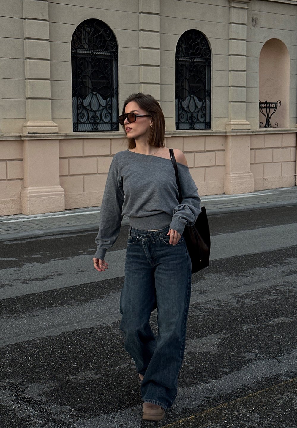 Donna con occhiali da sole, maglione grigio scivolato sulle spalle, jeans a gamba larga e una borsa nera che cammina su una strada lastricata vicino a un edificio beige.