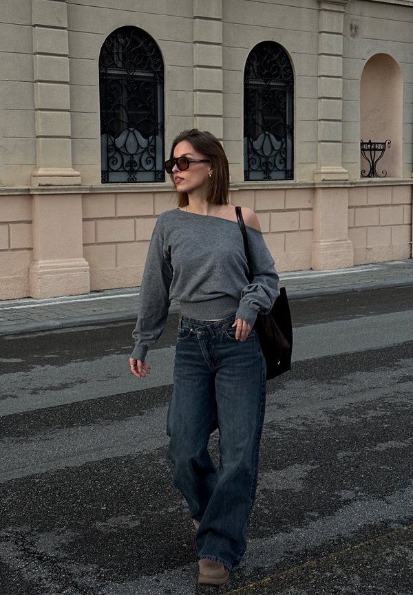 Nő napszemüvegben, szürke vállra húzott pulóverben, széles szárú farmerral és fekete táskát cipelve sétál egy burkolt utcán egy bézs épület mellett.