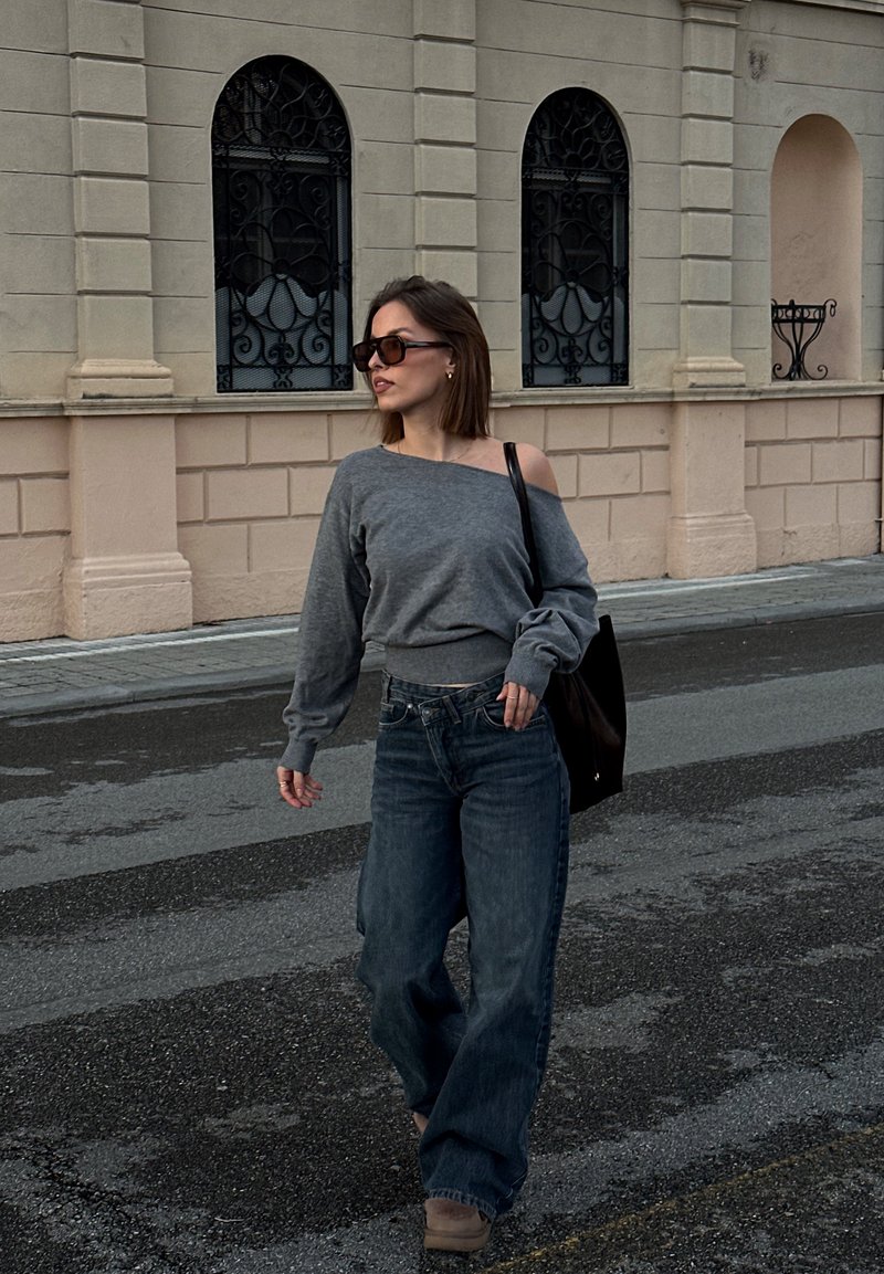 Frau mit Sonnenbrille, grauem Schulterfrei-Pullover, weit geschnittenen Jeans und einer schwarzen Tasche, die auf einer gepflasterten Straße an einem beigefarbenen Gebäude entlangläuft.