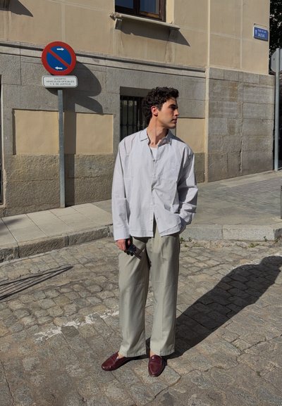 Joven con camisa de rayas claras y pantalones beige de pie en una calle adoquinada cerca de una señal de prohibido estacionar, mirando hacia la derecha y sosteniendo unas gafas de sol.