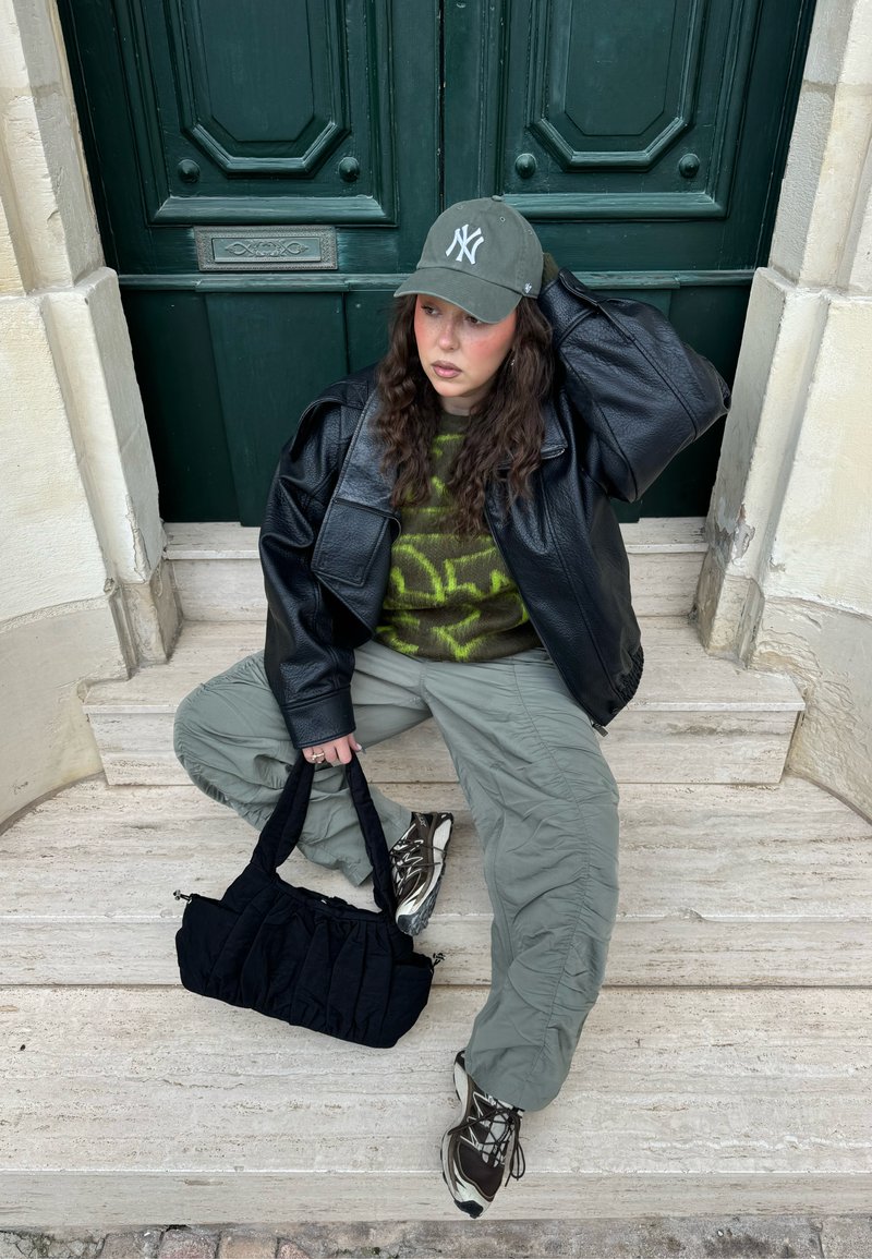 Young woman in green cap, black leather jacket, and cargo pants sitting on stone steps holding a black bag in front of dark green doors.