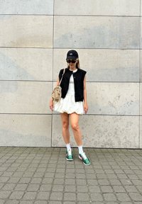 Svart ermeløs vest over en hvit topp, kombinert med et puffet hvitt skjørt. Grønn og rosa sneakers, hvite sokker og en svart cap. Beige veske.