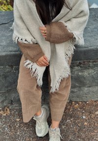 Châle gris clair à franges avec une texture douce, porté sur un pull marron en tricot et assorti à un pantalon en velours côtelé beige et des baskets grises.