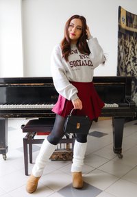 Felpa grigia con testo bordeaux, gonna a pieghe bordeaux, borsa nera, collant neri, scaldamuscoli bianchi e stivaletti beige, posata accanto a un pianoforte.