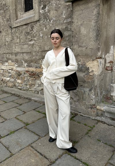 Cardigan de gran tamaño en crema, pantalones anchos blancos y bolso negro. El conjunto presenta un escote en forma de V, un corte relajado y texturas suaves. Fondo de pared de piedra.