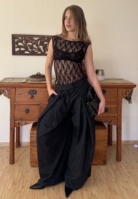 Body noir en dentelle avec manches courtes, associé à un pantalon ample noir à fines rayures. Le mannequin tient un petit sac à main noir, debout près d'un meuble en bois.