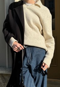 Μπεζ, πλεκτό πουλόβερ κάτω από ένα μαύρο παλτό; σκούρα τζιν παντελόνια. Τα αξεσουάρ περιλαμβάνουν ένα ασημένιο δαχτυλίδι και μαύρα γυαλιά ηλίου. Υφές υλικών.