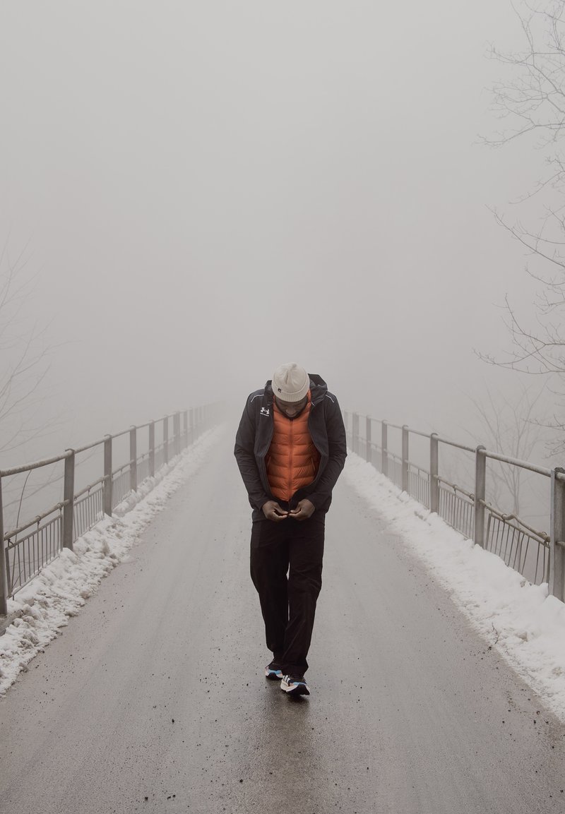 Man die een oranje vest en een witte muts draagt, loopt alleen over een besneeuwde brug in dichte mist en kijkt naar zijn telefoon in zijn handen.