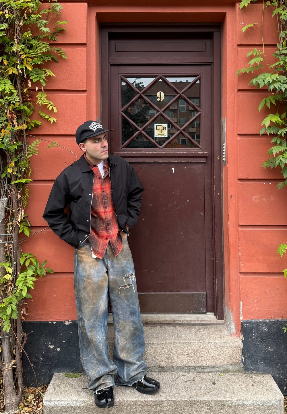Homme en veste noire, chemise à carreaux rouge, jean bleu usé et casquette, debout sur des marches devant une porte brune avec des vignes sur un mur orange.