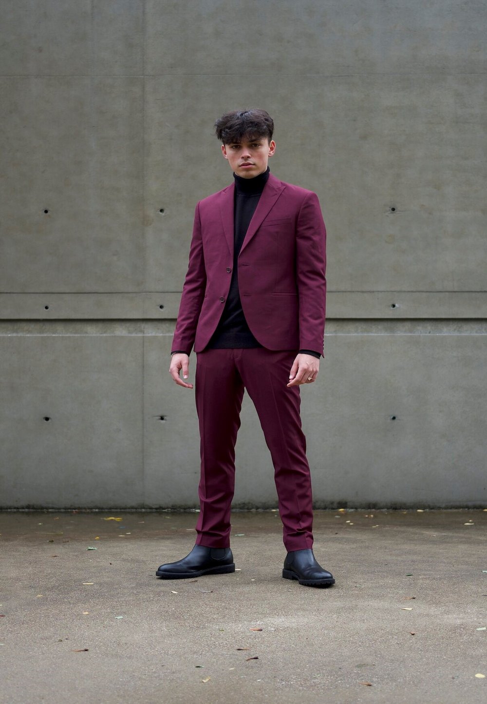 Burgunderfarget dress med en tettsittende blazer og matchende bukser, kombinert med en svart høyhalset genser. Støvlene er svarte, glatte og ankellange. Betongbakgrunn.