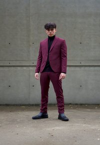 Burgunderfarget dress med en tettsittende blazer og matchende bukser, kombinert med en svart høyhalset genser. Støvlene er svarte, glatte og ankellange. Betongbakgrunn.