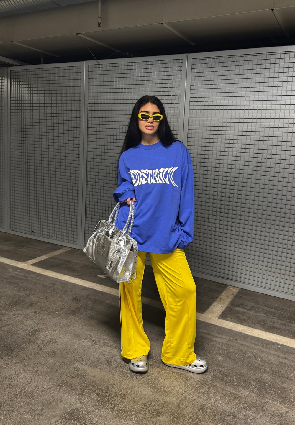 Sweatshirt azul oversized com gráfico branco, calças largas amarelas, handbag prateira, óculos de sol amarelos e calçado cinza. Fundo texturizado.