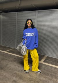 Sweatshirt azul oversized com gráfico branco, calças largas amarelas, handbag prateira, óculos de sol amarelos e calçado cinza. Fundo texturizado.