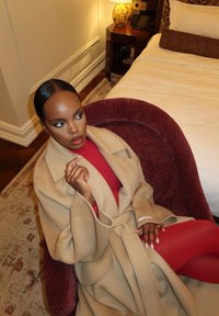 Un abrigo de lana color beige claro cubre un suéter de cuello alto rojo y medias rojas. La persona está sentada en una silla de felpa color burdeos con tela estampada.