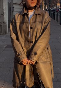 Απαλή καφέ υπερμεγέθης καπαρντίνα με γυαλιστερή φινίρισμα, διαθέτει μαύρα κουμπιά, μεγάλες τσέπες και λεπτομέρειες ραφής.