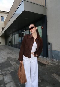 Femme portant une veste marron, un haut blanc et un pantalon blanc, tenant un sac marron, debout devant un bâtiment moderne aux fenêtres en verre.