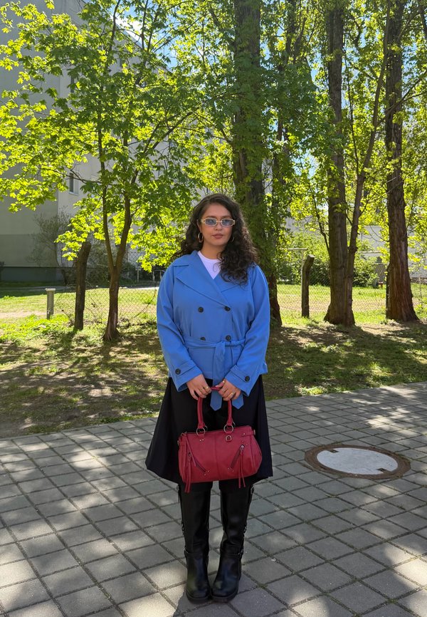 Jeune femme aux longs cheveux bouclés portant des lunettes de soleil, un manteau bleu, une jupe noire et des bottes, tenant un sac à main rouge à l'extérieur près des arbres.