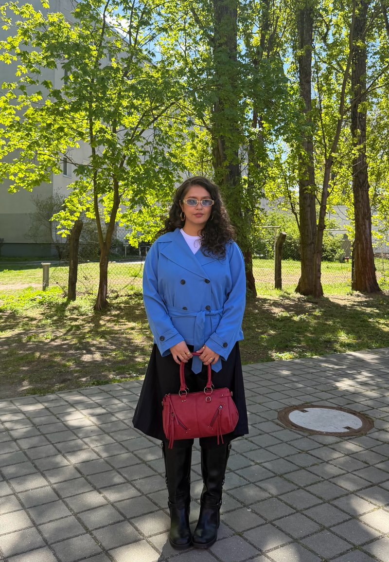 Jeune femme aux longs cheveux bouclés portant des lunettes de soleil, un manteau bleu, une jupe noire et des bottes, tenant un sac à main rouge à l'extérieur près des arbres.