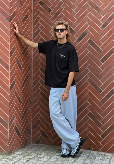 Camiseta negra de gran tamaño con logo blanco, vaqueros holgados de color azul claro, zapatillas negras y blancas con estampado, de pie contra una pared de ladrillos en espiga.