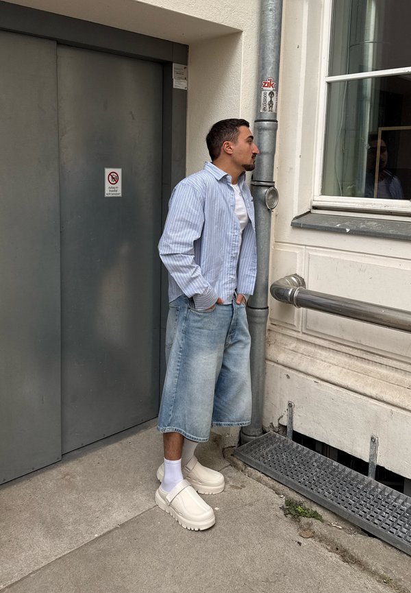 Homme en chemise rayée et short en denim jusqu’aux genoux, debout, les mains dans les poches, sur le trottoir de la ville près d’une porte grise et d’une fenêtre.
