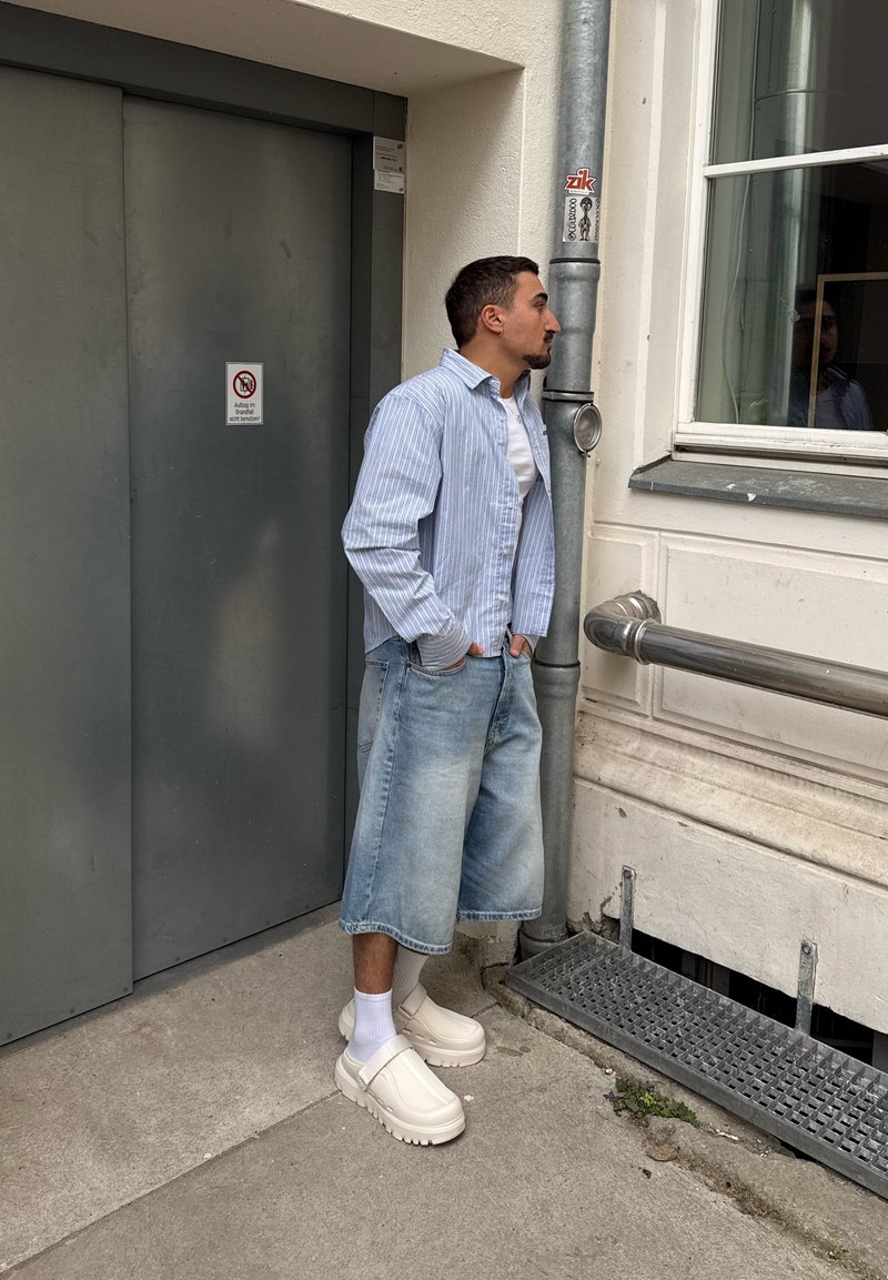Homme en chemise rayée et short en denim jusqu’aux genoux, debout, les mains dans les poches, sur le trottoir de la ville près d’une porte grise et d’une fenêtre.