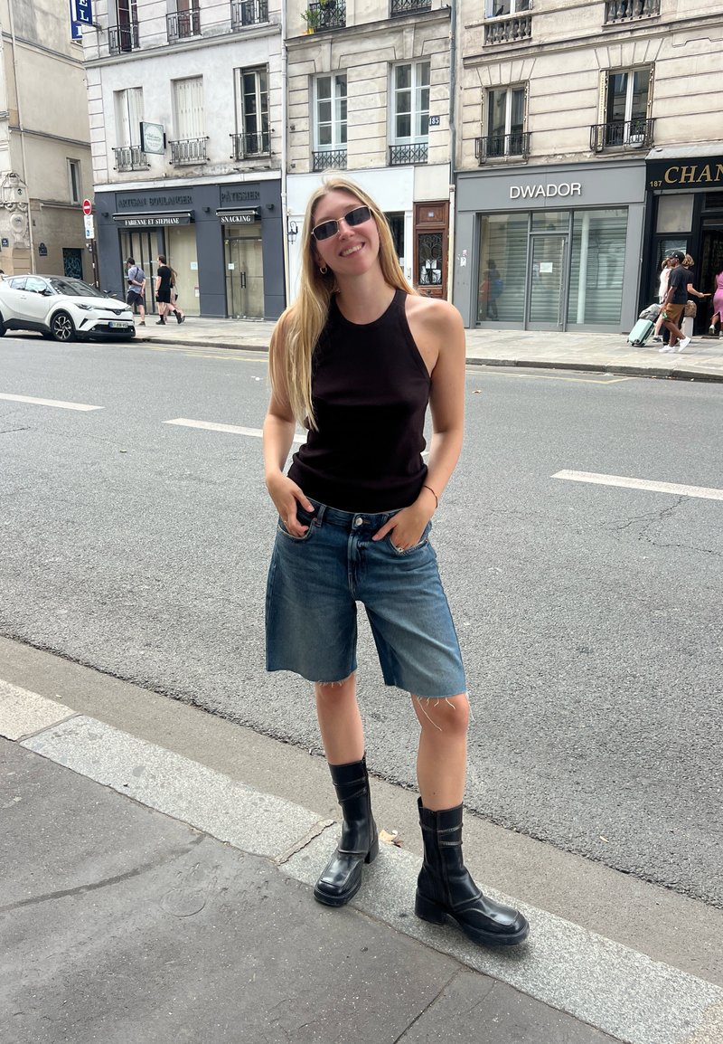 Μαύρη αμάνικη μπλούζα, μπλε τζιν σορτς με ξέφτια, μαύρες μπότες μέχρι τον αστράγαλο, στέκεται σε έναν δρόμο με αστικό φόντο.