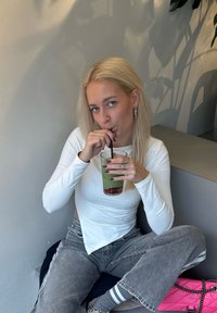 Prozoren plastični kozarec z zeleno pijačo in rdečim sadjem, ženska v beli dolgi majici, sivih širokih hlačah, drži slamico, sproščeno notranje okolje.