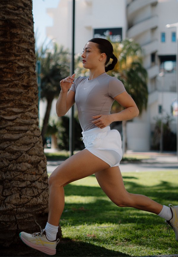 Jeune femme faisant du jogging en plein air près d'un palmier, portant un t-shirt gris, un short blanc et des baskets, avec des bâtiments urbains en arrière-plan.