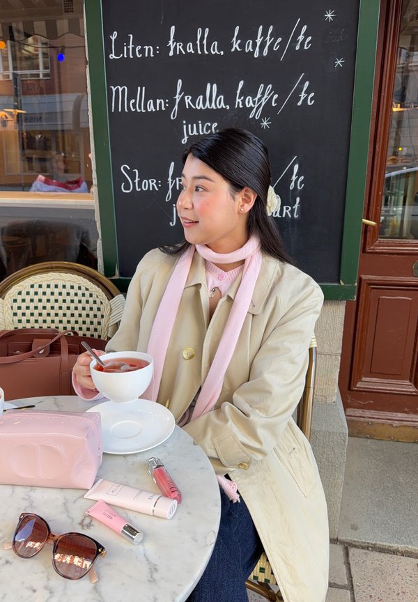 Femme en manteau beige et écharpe rose tenant une tasse de thé à une table de café, avec des produits de beauté, des lunettes de soleil et un tableau de menu derrière elle.